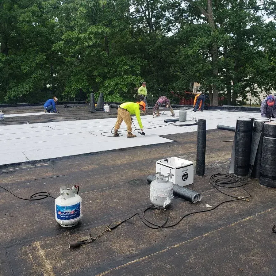 Roofing crew rolling light-colored sheets over a black base layer on a large flat roof, with rolls of roofing material and propane tanks