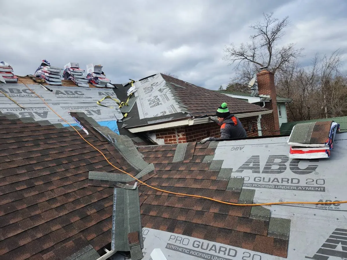 Roofer installing brown architectural shingles over gray synthetic underlayment, with shingle bundles stacked along the ridge