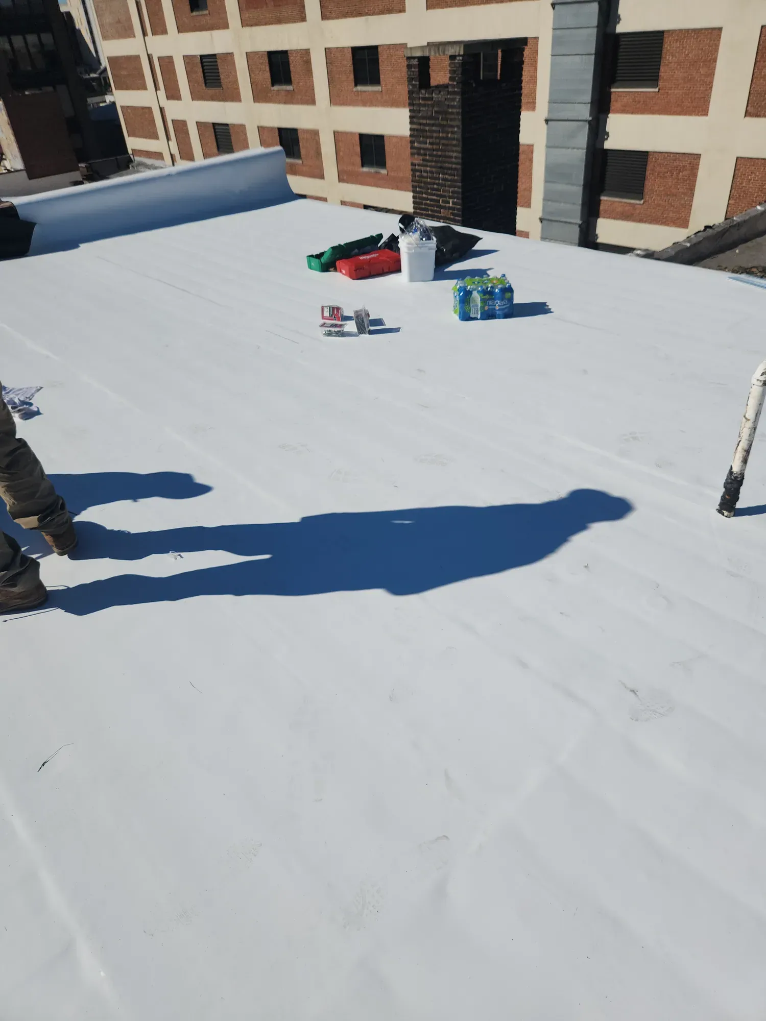 White single-ply membrane on a flat roof with toolboxes and fasteners, next to a brick chimney and a tall brick building